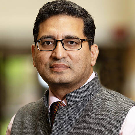 A man wearing glasses, a pink shirt, patterned tie, and a gray vest looks directly at the camera.