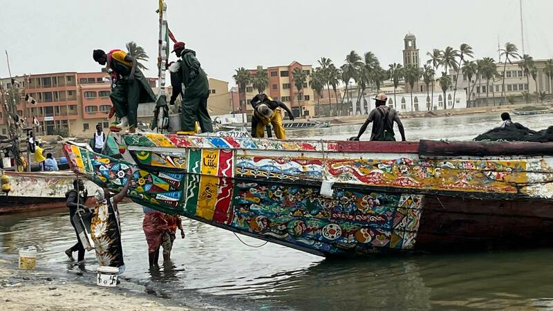 A heavily decorated boat is coming into shore in Senegal with 5 men on the boat and 4 on shore greeting the boat to unload it.