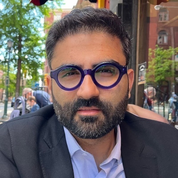 Headshot of a person with dark hair, beard, and distinctive round, purple eyeglasses, wearing a dark blazer and light blue collared shirt.