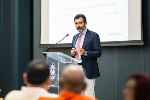 Bearded man in navy suit and pink tie, Reza Baqir, speaks passionately at a transparent podium with the Notre Dame seal.