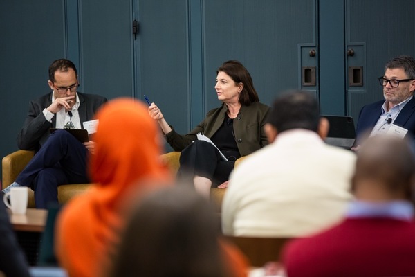 Panelists sit in yellow chairs against a teal wall. Woman in olive jacket, Ana Maria Ibanez, gestures with a pen; man (Cesar Sosa-Padilla) on left thoughtful, man on right (Kevin Gallagher) listens.