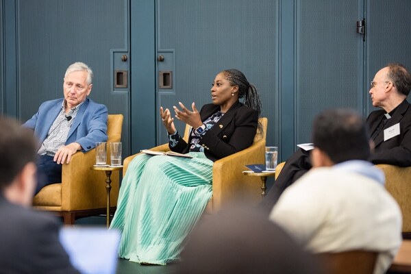 Three panelists in armchairs. A woman in a black blazer and green skirt gestures while speaking. A man in a blue blazer and another in clerical attire listen.